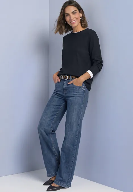 Frau in schwarzem Pullover und weit geschnittenen Jeans posiert lächelnd mit Händen in den Taschen.