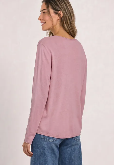 Eine Frau in rosa Langarmshirt und blauen Jeans steht seitlich, lächelt.