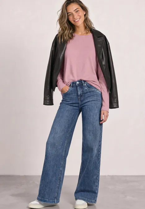 Lächelnde Frau in rosa Pullover, weiten Jeans und schwarzer Lederjacke vor neutralem Hintergrund.