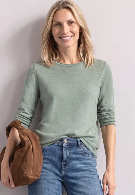 Trui met ronde hals en omgeslagen randen eucalyptus green mel. Een lachende blonde vrouw in een groene trui en spijkerbroek, die een bruine jas vasthoudt.