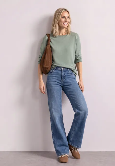 Trui met ronde hals en omgeslagen randen eucalyptus green mel. Een glimlachende vrouw in groene trui, wijde high-waist jeans, met bruine schoudertas en schoenen.