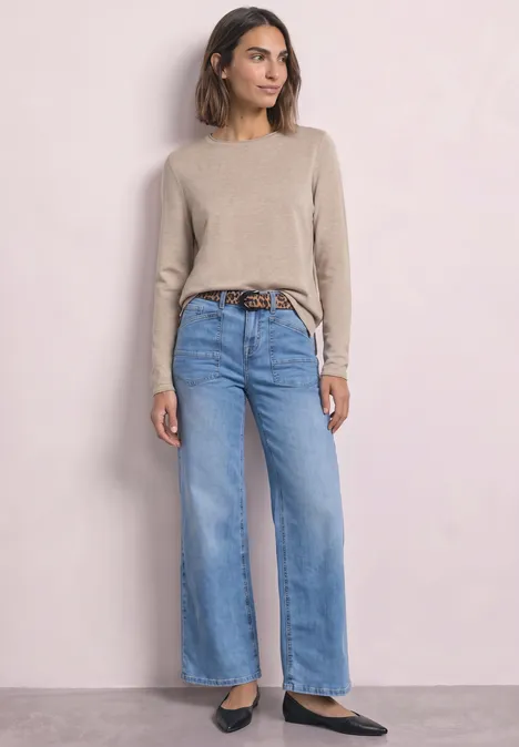 Trui met ronde hals en omgeslagen randen clay sand mel. Vrouw in beige trui met wijde lichtblauwe jeans, bruine riem en zwarte schoenen voor een lichte achtergrond.