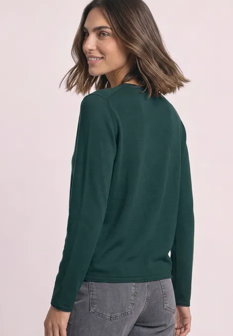 Trui met ronde hals en omgeslagen randen shadow teal Jonge vrouw met halflang bruin haar lacht over haar schouder en draagt een donkergroene trui met grijze jeans.