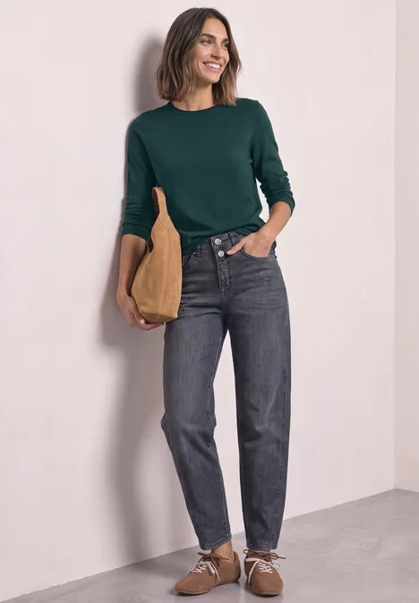 Trui met ronde hals en omgeslagen randen shadow teal Een glimlachende vrouw in een groen shirt en spijkerbroek leunt tegen een muur en houdt een beige tas vast.