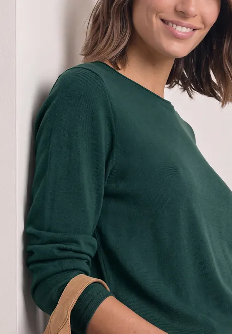 Trui met ronde hals en omgeslagen randen shadow teal Een vrouw leunt tegen een muur, draagt een groen shirt met lange mouwen en glimlacht zacht.