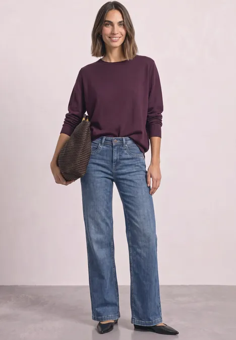 Trui met ronde hals en omgeslagen randen berry maroon Een glimlachende vrouw in paarse trui, wijde jeans, witte sneakers en tas, staand voor een lichte achtergrond.