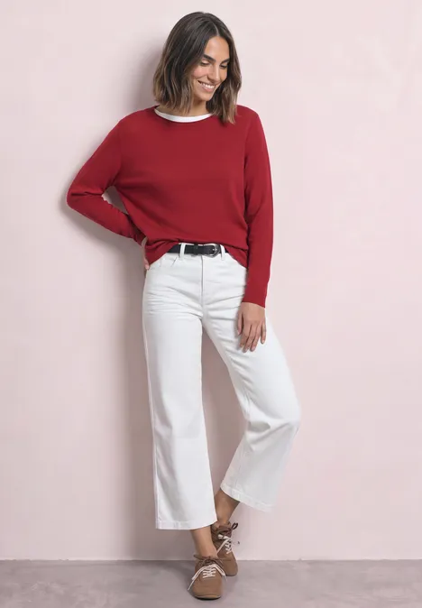 Trui met ronde hals en omgeslagen randen inferno red Vrouw in rode trui, witte wijde cropped jeans en bruine sandalen tegen een roze achtergrond.