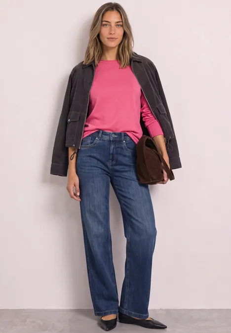 Trui met ronde hals en omgeslagen randen impact pink Vrouw staand met een roze top, zwarte jas, wijde blauwe jeans, zwarte schoenen en een bruine tas.