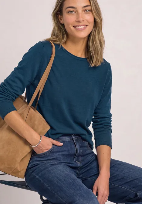 Frau in tealfarbenem Langarmshirt und Jeans sitzt lässig mit brauner Wildledertasche und lächelt.