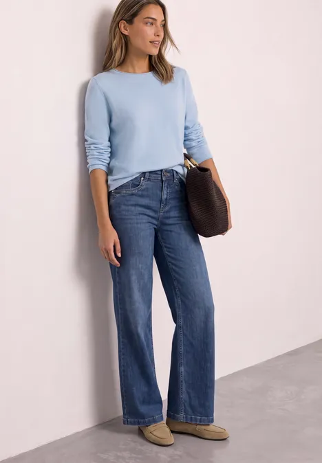 Trui met ronde hals en omgeslagen randen pale blue mel. Vrouw in lichtblauwe trui, donker wijde jeans en pantoffels leunt nonchalant tegen een lichte muur met clutch.