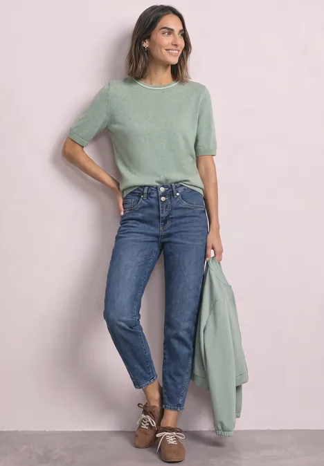 Frau trägt grünes T-Shirt, blaue Jeans, braune Schuhe, hält passendes Strickjacke in der Hand, steht vor rosa Wand.