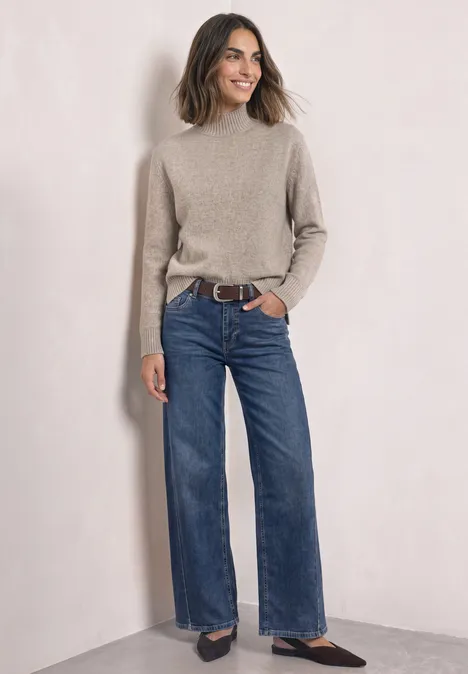 Femme debout devant un mur clair, portant un pull beige, un jean large et des chaussures foncées.