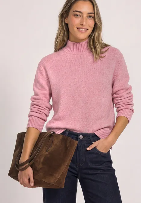 Femme portant un pull rose et un jean foncé, tenant un sac en daim marron, elle sourit.