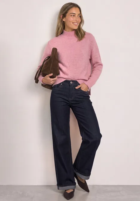 Femme en pull rose, jean ample bleu et chaussures marron, portant un sac à main et souriant devant un mur clair.