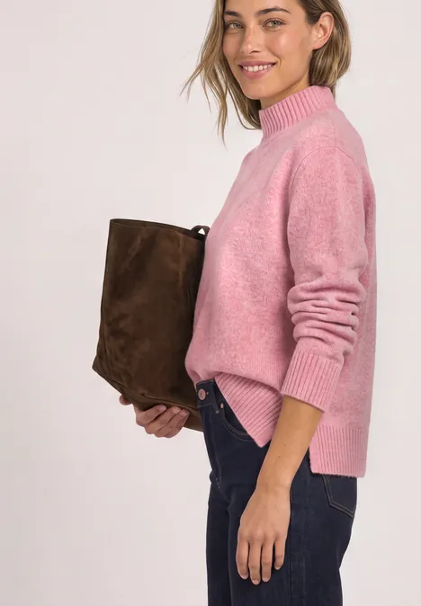 Femme en pull rose et jean, tenant un sac marron en daim, souriante de profil.