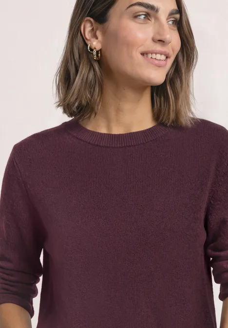 Femme aux cheveux bruns, souriante, porte un pull violet foncé et des créoles dorées.