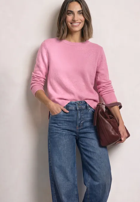 Une femme souriante porte un pull rose, un jean et tient un sac bordeaux devant un fond clair.