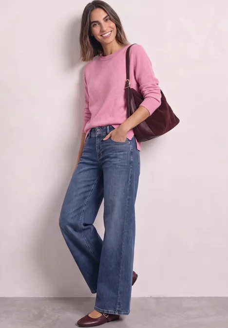 Jeune femme souriante vêtue d'un jean ample et d'un pull rose, portant un sac à épaule marron.