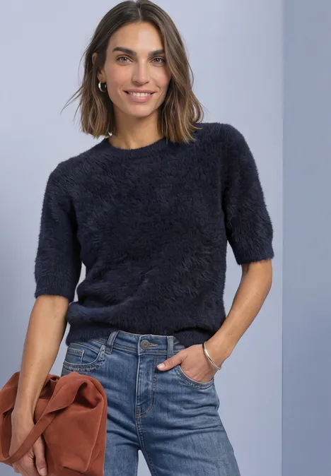 Frau in dunkelblauem, flauschigem Kurzarm-Pullover und Jeans, lächelt, hält braune Tasche, vor hellblauem Hintergrund.