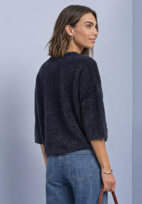 Frau von der Seite, trägt schwarzen, flauschigen Pullover mit 3/4-Ärmeln und blaue Jeans.