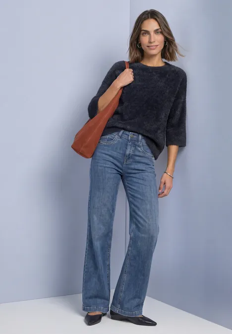 Frau mit dunkelgrauem Pullover, hoch taillierten Schlagjeans, brauner Schultertasche und schwarzen Schuhen vor neutralem Hintergrund.