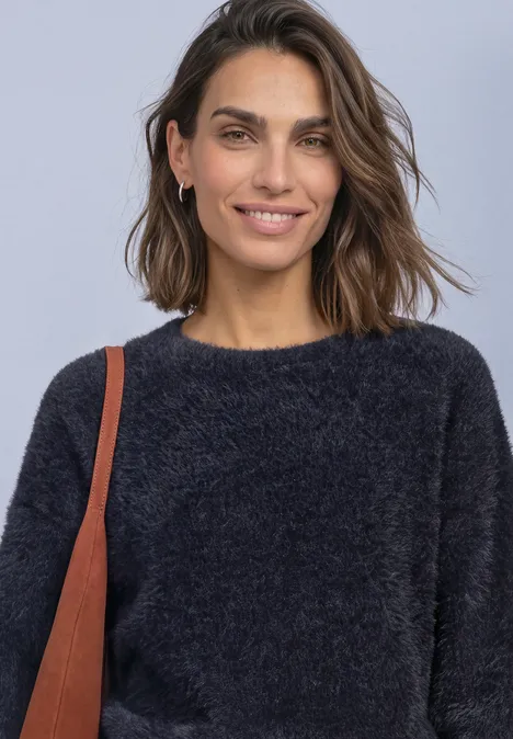 Eine Frau mit kurzen braunen Haaren, lächelt, trägt einen flauschigen schwarzen Pullover und eine Umhängetasche.
