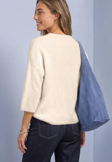 Femme en pull crème, manches 3/4, porte un sac bleu sur l’épaule, vue de côté regardant à gauche.
