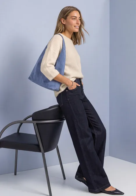 Femme en jean large, haut beige et bleu clair, avec sac bleu, près d'une chaise sur fond minimaliste.
