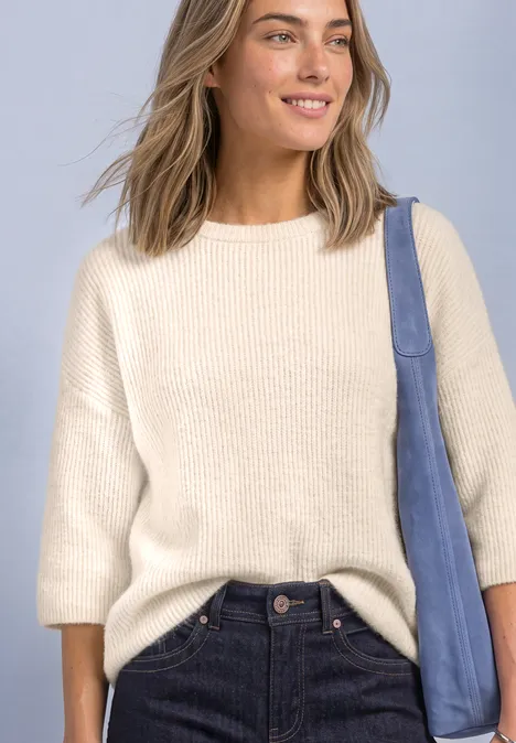 Femme souriante en pull crème à manches 3/4, jean foncé et sac à épaule bleu devant un fond clair.