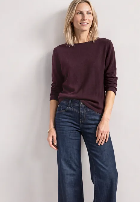 Vrouw in bordeauxrode trui en wijde jeans, poserend voor een neutrale achtergrond.