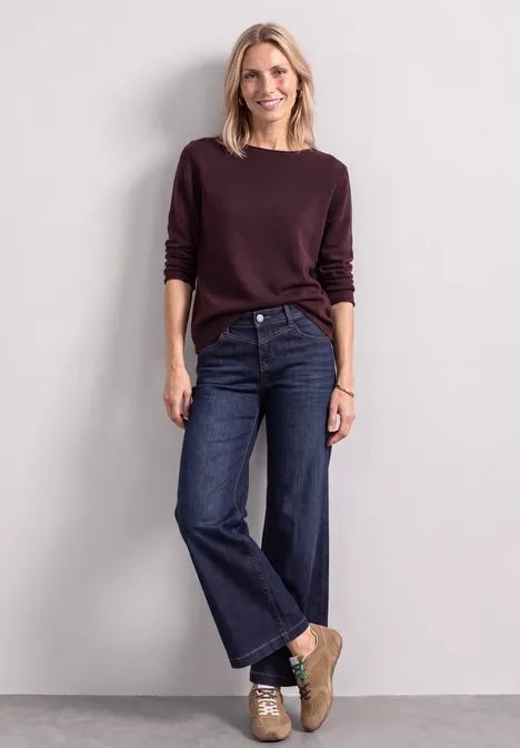 Een vrouw met blond haar draagt een bordeaux trui, wijde jeans en sneakers voor een neutrale achtergrond.