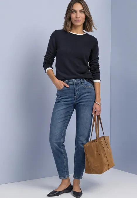 Frau in schwarzem Pullover, blauen Jeans und schwarzen Loafers, hält eine braune Tasche.