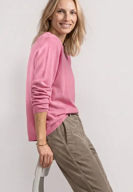 Frau im rosa Pullover und beigefarbenen Hosen, seitlich stehend, lächelnd vor hellem Hintergrund.