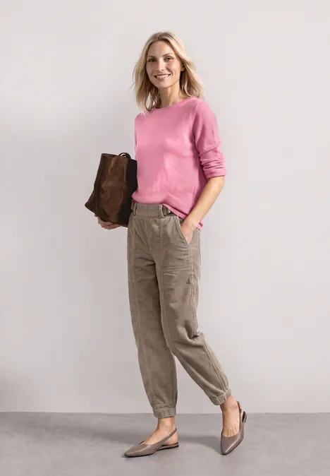 Frau in rosa Pullover, khakifarbener Cargo-Hose und brauner Tasche steht vor hellem Hintergrund.