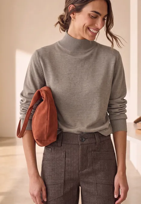 Basic Pullover mit Turtle Neck sanded amber mel. Frau mit grauem Rollkragenpullover und braunen Hosen, lächelt und trägt eine rostrote Jacke über dem Arm.