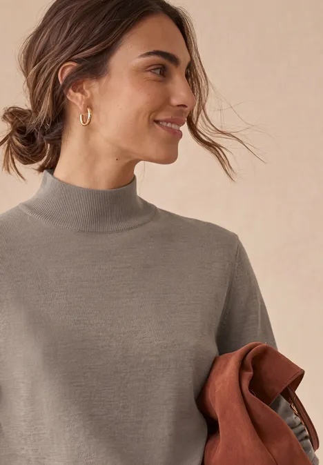 Basic Pullover mit Turtle Neck sanded amber mel. Junge Frau im grauen Rollkragenpullover hält eine braune Jacke, lächelt seitlich vor hellem Hintergrund.