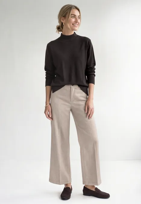 Frau vor weißem Hintergrund in schwarzem Longsleeve, beiger weiter Hose und braunen Loafern.