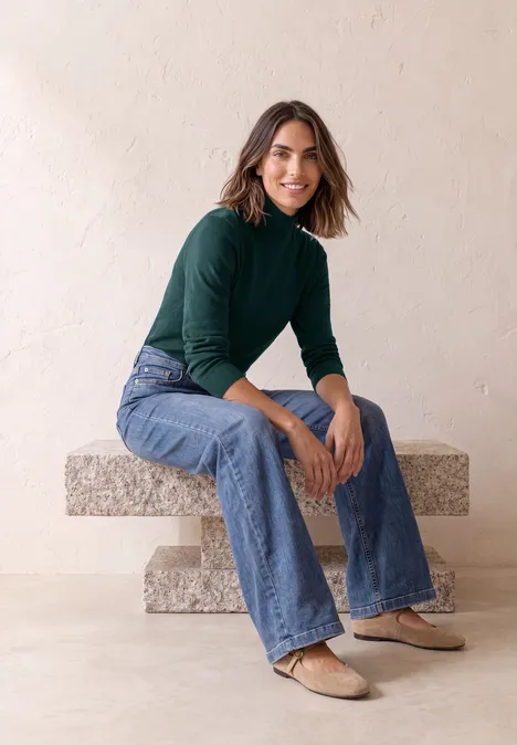 Basic Pullover mit Turtle Neck shadow teal Eine Frau sitzt auf einer Steinbank, trägt grünen Rollkragenpullover, Jeans und Ballerinas.