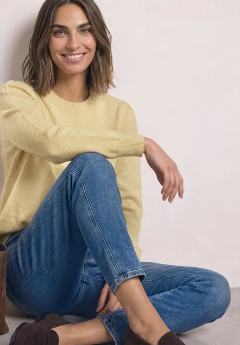 Frau in gelbem Pullover und blauer Jeans sitzt entspannt und lächelt.