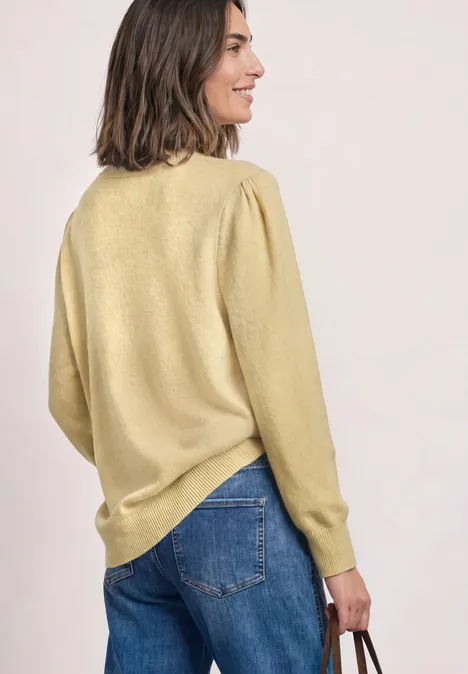 Frau in gelbem Pullover und Jeans, seitlich vom Rücken fotografiert, hält eine Tasche in der rechten Hand.
