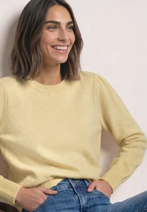 Frau im pastellgelben Pullover und blauer Jeans sitzt locker und lächelt.