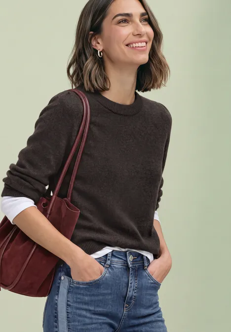 Junge Frau mit braunem Pullover über weißem Shirt, Jeans und roter Schultertasche lächelt.