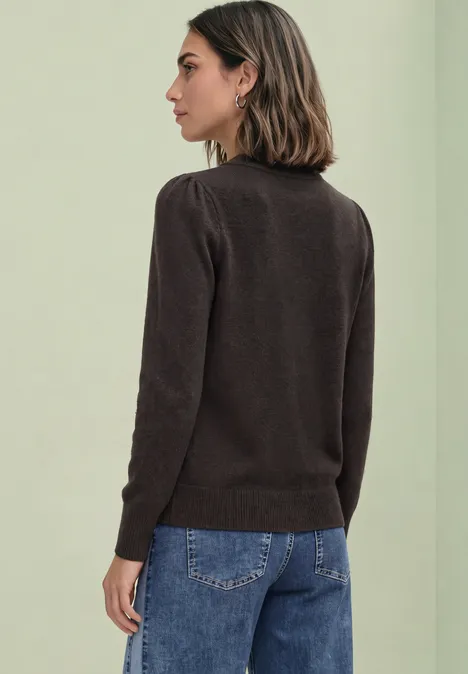 Frau im Profil trägt braunen Pullover und Jeans vor schlichtem, hellem Hintergrund.