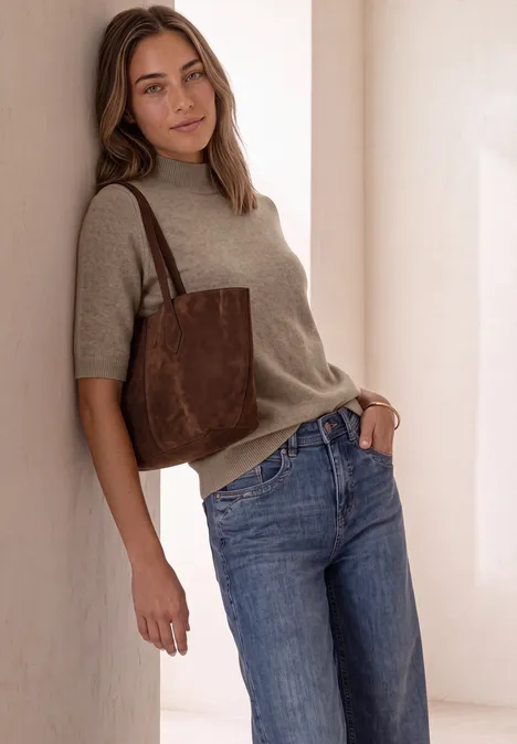 Kurzarm Pullover mit Turtle Neck sanded amber mel. Junge Frau lehnt an heller Wand, trägt beigefarbenen Pullover, blaue Jeans und braune Schultertasche.