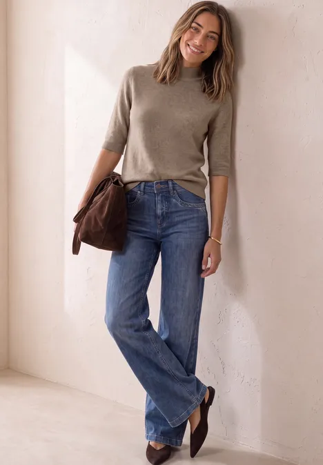 Kurzarm Pullover mit Turtle Neck sanded amber mel. Eine lächelnde Frau in beigefarbenem Pullover und blauen Jeans lehnt entspannt an einer Wand, trägt Tasche.