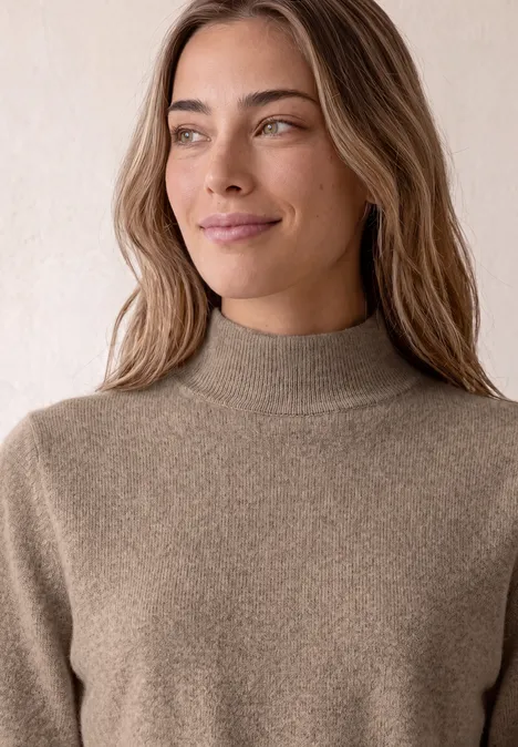 Kurzarm Pullover mit Turtle Neck sanded amber mel. Junge Frau mit langem, gewelltem Haar trägt einen hellbraunen Rollkragenpullover und blickt lächelnd zur Seite.