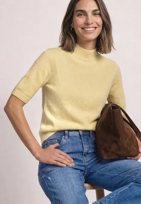 Eine lächelnde Frau sitzt auf einem Hocker, trägt einen gelben Kurzarmpullover, blaue Jeans und hält eine braune Tasche.