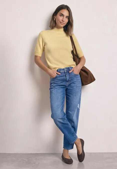 Eine Frau in gelbem Shirt, blauen Jeans und brauner Tasche vor rosa Hintergrund.