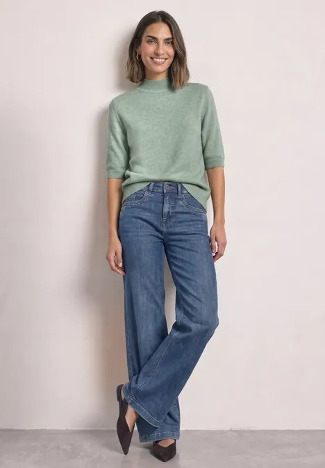 Eine Frau mit grünem Pullover, weitem Jeans und schwarzen Schuhen lehnt lächelnd an einer hellen Wand.