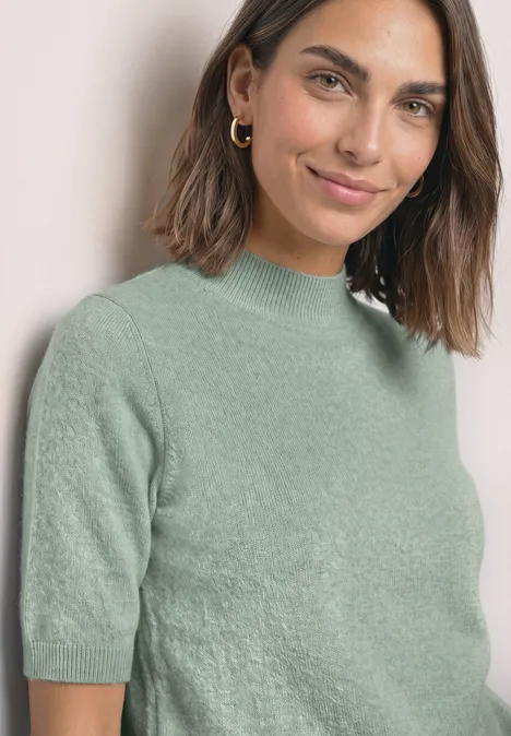 Eine lächelnde Frau mit schulterlangen dunklen Haaren in einem kurzärmeligen, hellgrünen Pullover.